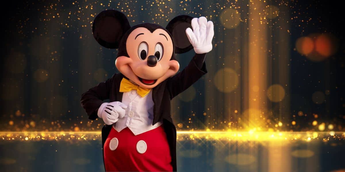 A person in a Mickey Mouse costume is waving and smiling. Mickey Mouse wears his iconic red shorts, yellow bow tie, and white gloves, standing against a sparkling, golden background at a Disney Park.