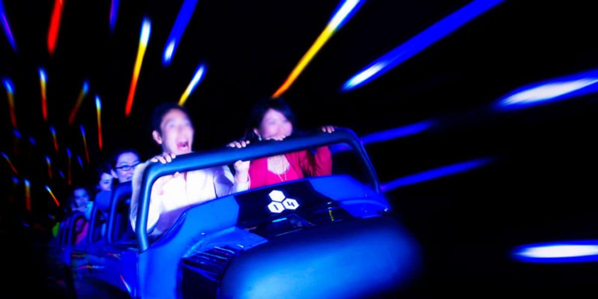 A group of people are riding a roller coaster in a dark environment with colorful light streaks creating a sense of motion and excitement. The passengers appear thrilled, with some screaming and others smiling as they hold onto the safety bar, enjoying the iconic Disney experience on Space Mountain.