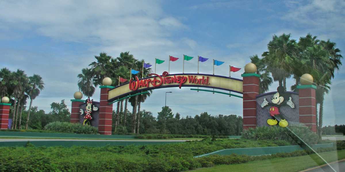 The entrance sign to Disney World showcases Mickey and Minnie Mouse. The archway, highlighted by colorful flags, stands proudly against a backdrop of palm trees and a cloudy sky, perfectly capturing the enchanting spirit of Walt Disney World.