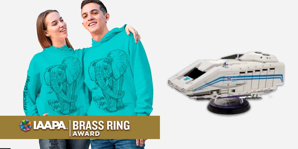 Two people wear matching turquoise hoodies with elephant designs, standing next to a futuristic white ride vehicle model. A gold banner at the bottom reads "IAAPA Brass Ring Award.