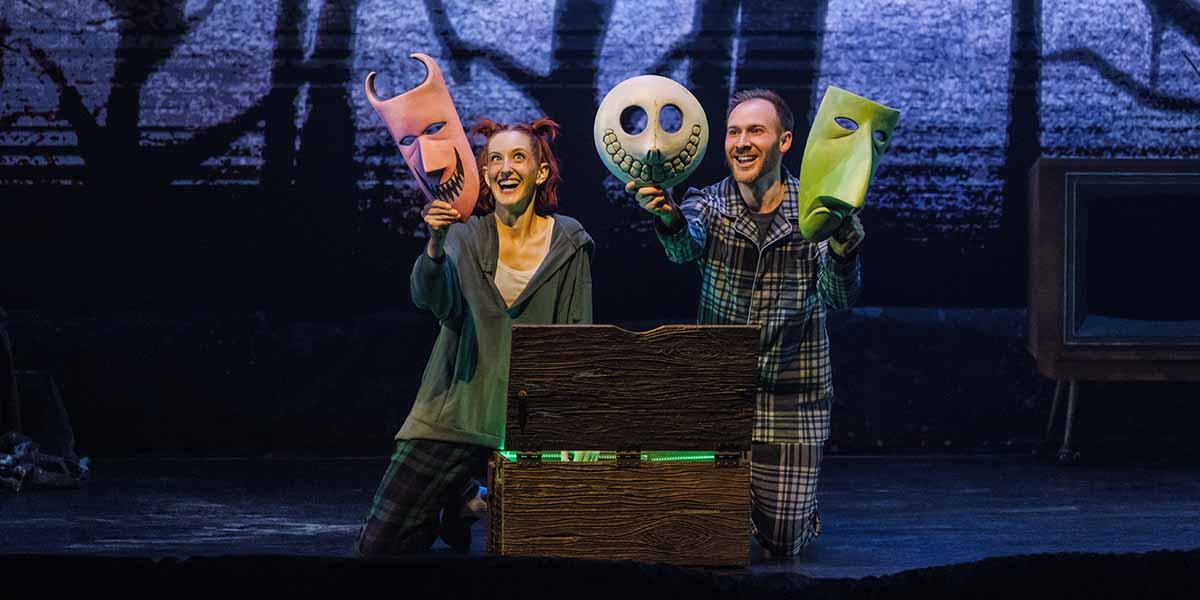 Two performers in pajamas kneel on stage, smiling and holding up large colorful masks—a pink devil, a white skull, and a green face—beside an open wooden chest, with a dark, forest-like backdrop behind them.