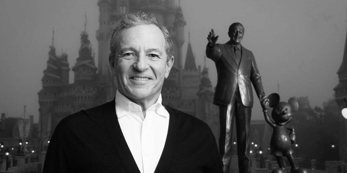 A man smiles broadly while standing in front of a castle and a statue of a man holding hands with a mouse character, iconic symbols of the Disney Company. The image is in black and white, and the atmosphere appears slightly foggy.