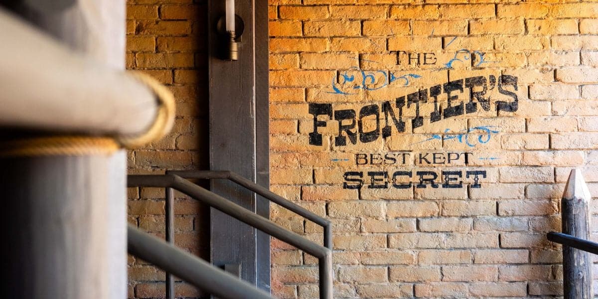 A brick wall with black and blue text reading "The Frontier's Best Kept Secret" stands proudly as an opening day attraction. The wall, lined with metal railings and anchored by a large wooden pole on the left, exudes a rustic, old-world charm reminiscent of the Magic Kingdom.