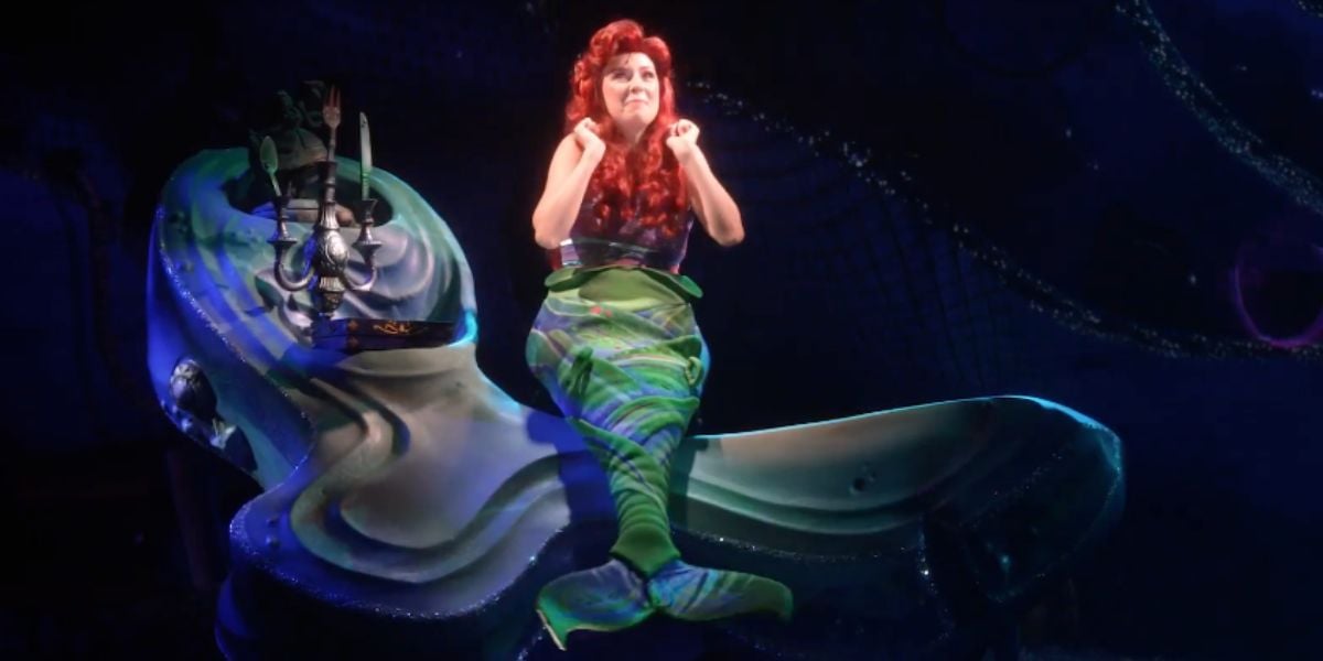 A performer dressed as Ariel from "The Little Mermaid" musical at Walt Disney World sits on a large seashell prop, wearing a red wig and green mermaid tail costume, smiling brightly under stage lights.