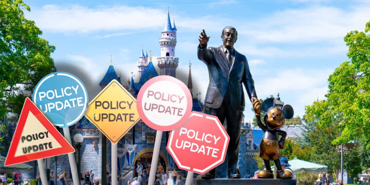 A statue of Walt Disney holding hands with a Mickey Mouse stands in front of a castle at a Disneyland Park in California. In the foreground, various road signs with "Policy Update" written on them are visible. The sky is clear and trees frame the image on the sides.