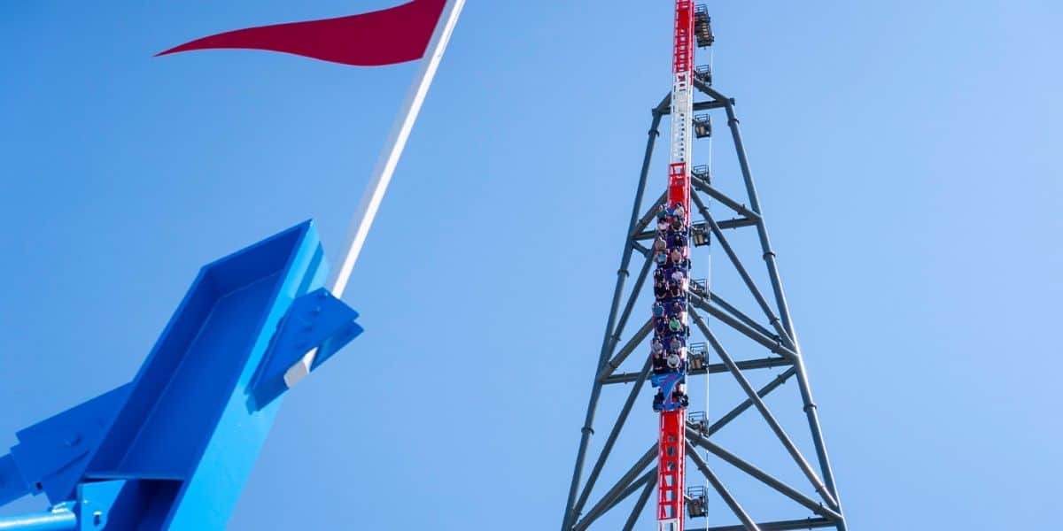 A group of people is ascending the tall, vertical roller coaster track of Top Thrill 2 with red and white sections at Cedar Point. A blue structure with a waving red flag is in the foreground, and the sky is clear and blue.