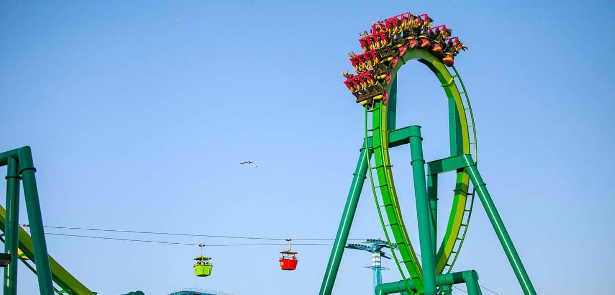 A vibrant green roller coaster at Cedar Point reaches its peak, looping upside down against a clear blue sky. Below, colorful gondolas—one red, one yellow—travel along a cable. A bird flies in the distance, creating an idyllic scene just before a sudden shutdown occurs.