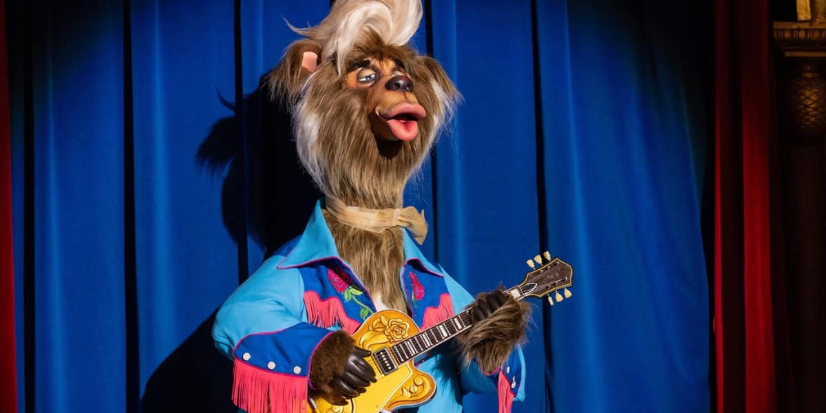 A bear-like animatronic character with light brown fur, a white tuft of hair on top, wearing a bright blue western-style jacket with red fringes, and holding a yellow guitar while standing in front of a blue curtain.