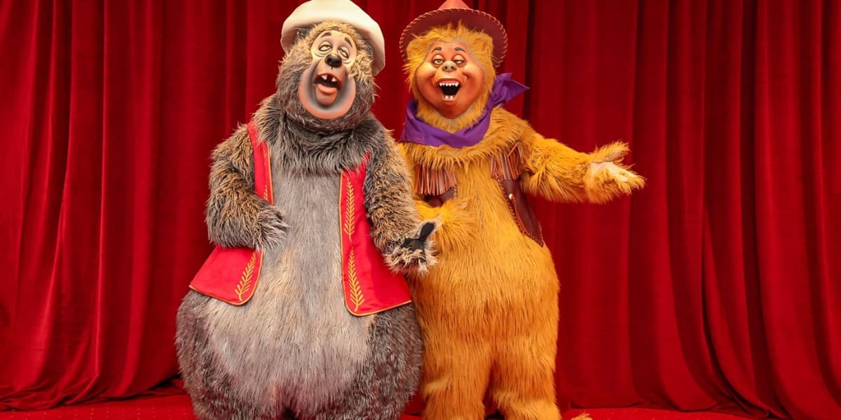 Two costumed characters stand side by side in front of a red curtain. The character on the left is a gray bear wearing a red vest and white hat, and the character on the right is a yellow bear wearing a cowboy outfit with a purple scarf. Both are smiling and singing.