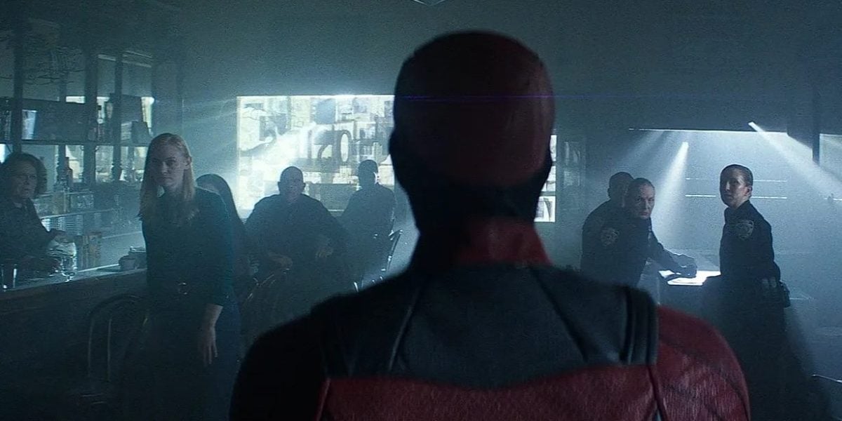 A man in a red superhero suit stands with his back to the camera, facing a group of people and police officers inside a dimly lit diner. Several people turn to look at him, appearing surprised or cautious.