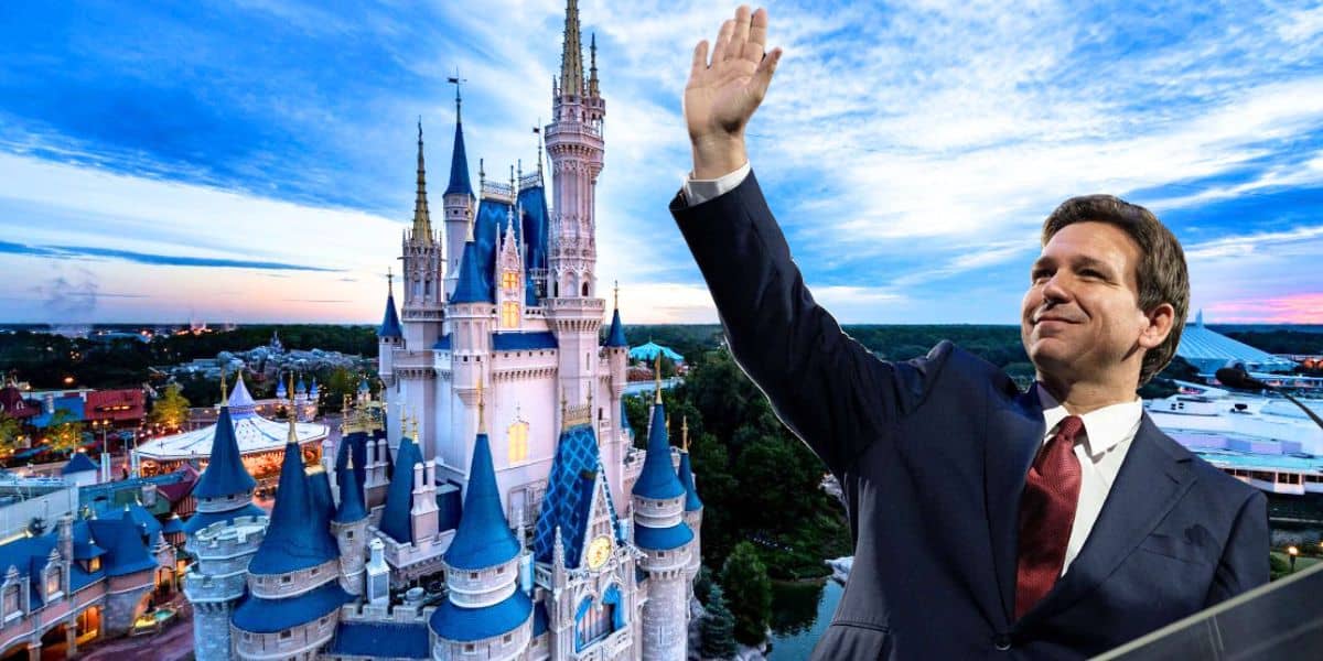 A man in a suit, with a red tie, is waving while standing in front of a tall, ornate castle reminiscent of a fantasy theme park. The sky is clear with light clouds. The background includes various buildings and attractions. DeSantis Disney World transportation expansion project.