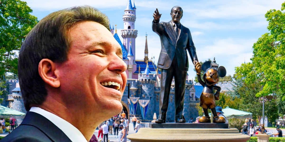 Ron DeSantis smiling in the foreground, with Sleeping Beauty Castle in Disneyland, a statue of Walt Disney holding Mickey Mouse's hand, and visitors enjoying the park visible in the background. The scene is bright and colorful, capturing a cheerful moment.