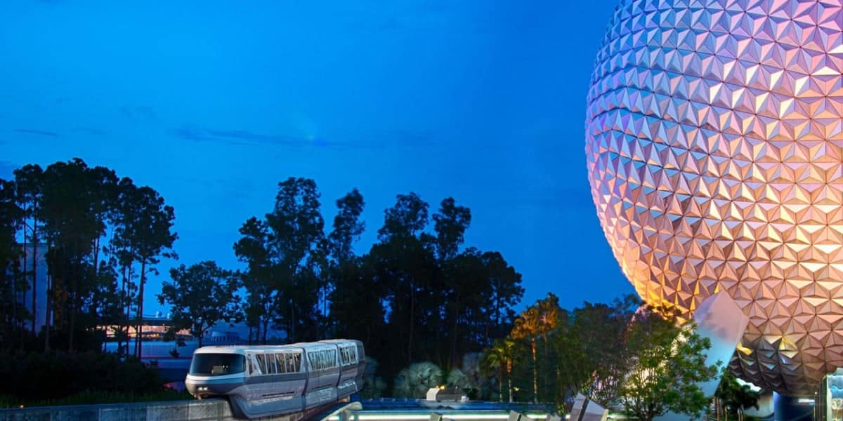 A monorail passes in front of a large geodesic sphere illuminated with purple and orange hues at dusk, with trees and a deep blue sky in the background.