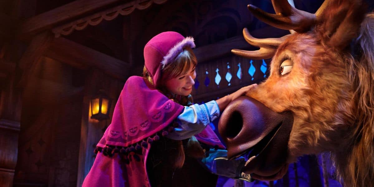 A young woman dressed in a pink winter cloak with a hood, adorned with black trim and designs, warmly pets a friendly reindeer with large antlers inside a cozy, wooden shelter illuminated by soft, warm light. She smiles as she interacts with the reindeer at the enchanting Disney park.