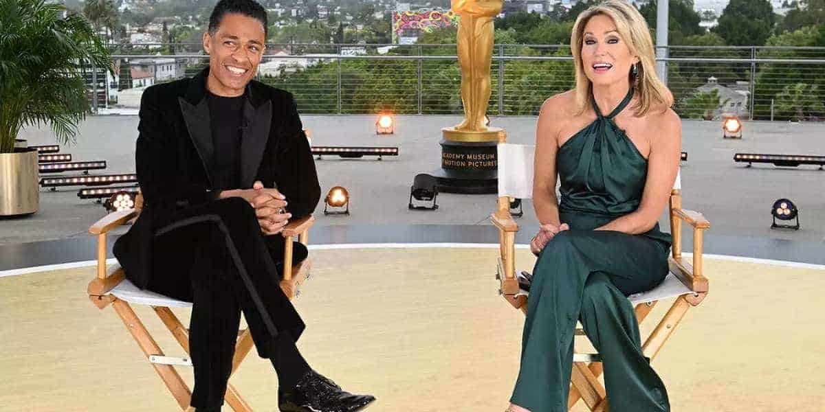 Amy Robach and TJ Holmes are seated on director's chairs on an outdoor terrace, with city buildings in the background and an Oscar statue behind them. TJ is wearing a black suit, while Amy stuns in a green jumpsuit. Both are smiling and appear to be hosting an event.