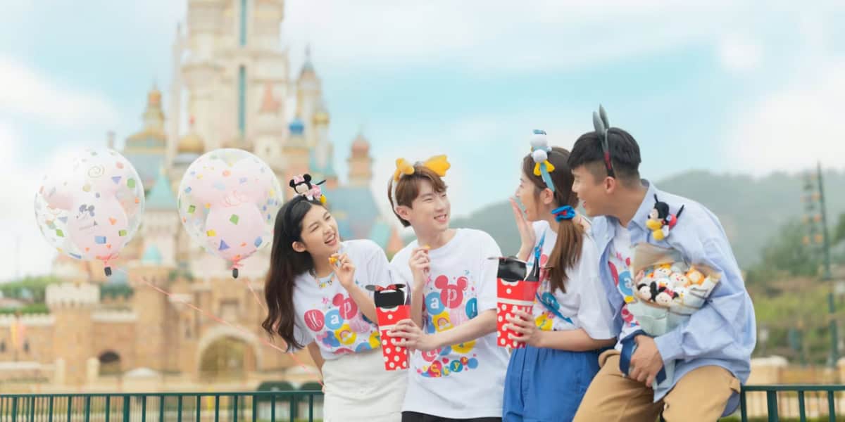 A group of four friends stands in front of a castle at a theme park. They are smiling and holding themed souvenirs, snacks, and balloons. Two of them wear headbands with cute characters, adding to the festive atmosphere. The sky is clear and sunny.