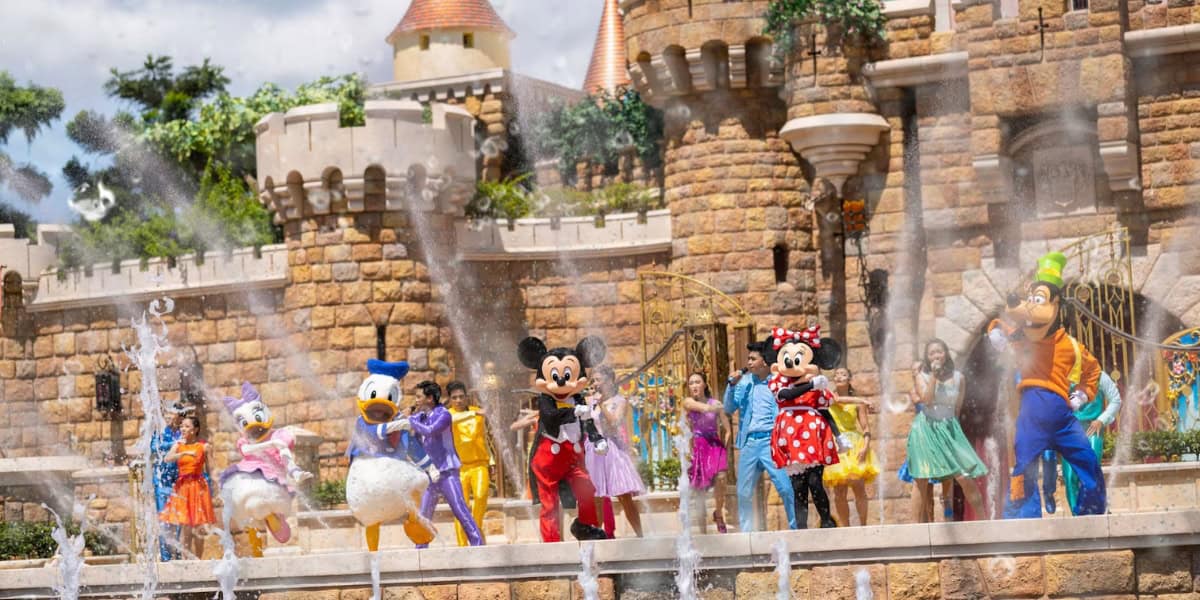 Disney characters, including Mickey Mouse, Minnie Mouse, Donald Duck, Daisy Duck, and Goofy, dance in front of a castle with water fountains spraying around them. Several other costumed characters are also performing on stage amidst a vibrant, festive atmosphere.
