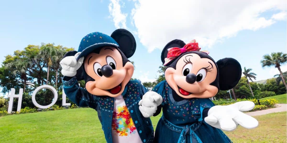 Mickey Mouse and Minnie Mouse, dressed in matching blue outfits with star patterns, pose cheerfully in an outdoor park setting. Trees and a blue sky with some clouds are visible in the background.