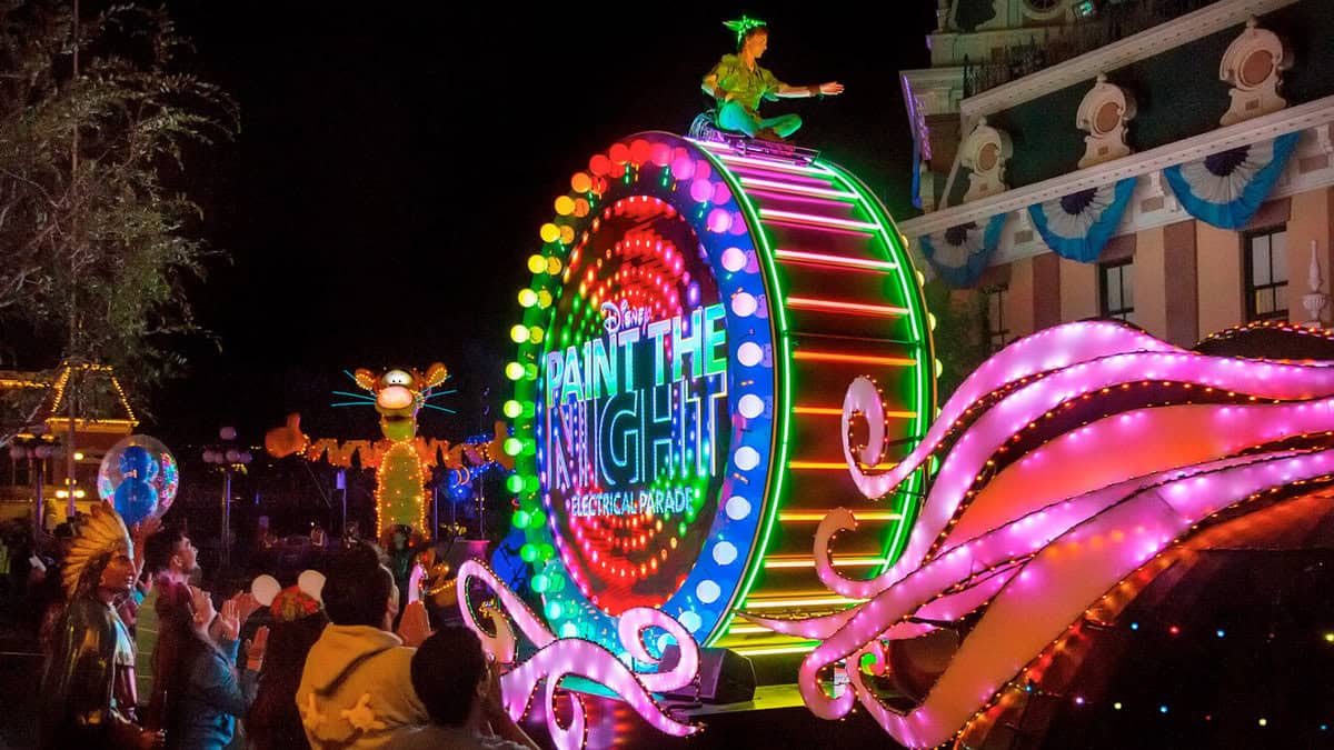 A vibrant nighttime Disneyland parade features a colorful, illuminated float with "Paint the Night" glowing brightly. A character is perched atop while neon figures dazzle in the background, captivating spectators watching from the sidelines or enjoying through a live stream.
