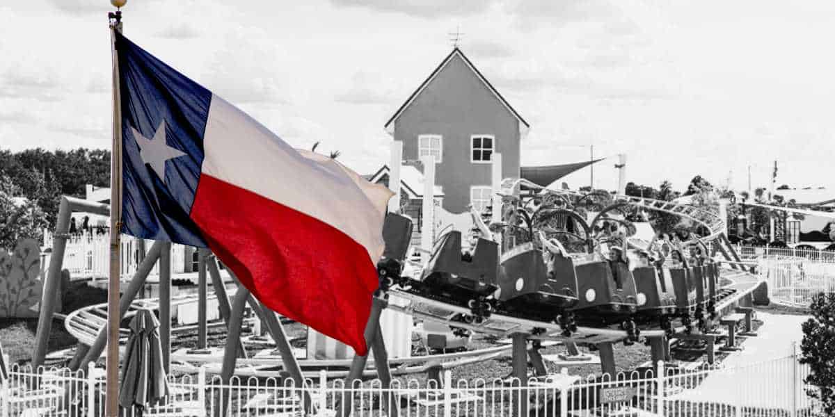 A Texas flag waves prominently in the foreground of a brand-new theme park scene. In the background, a red barn-like building and a roller coaster with several cars carrying passengers can be seen. The image is largely black and white, except for the flag and coaster cars.