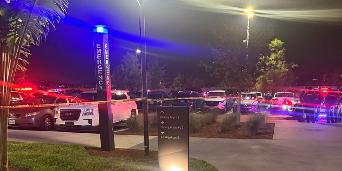 A parking lot at night with police cars and emergency vehicles flashing red and blue lights after a late-night shooting. Yellow caution tape blocks off the area, and an emergency call station is visible in the foreground.