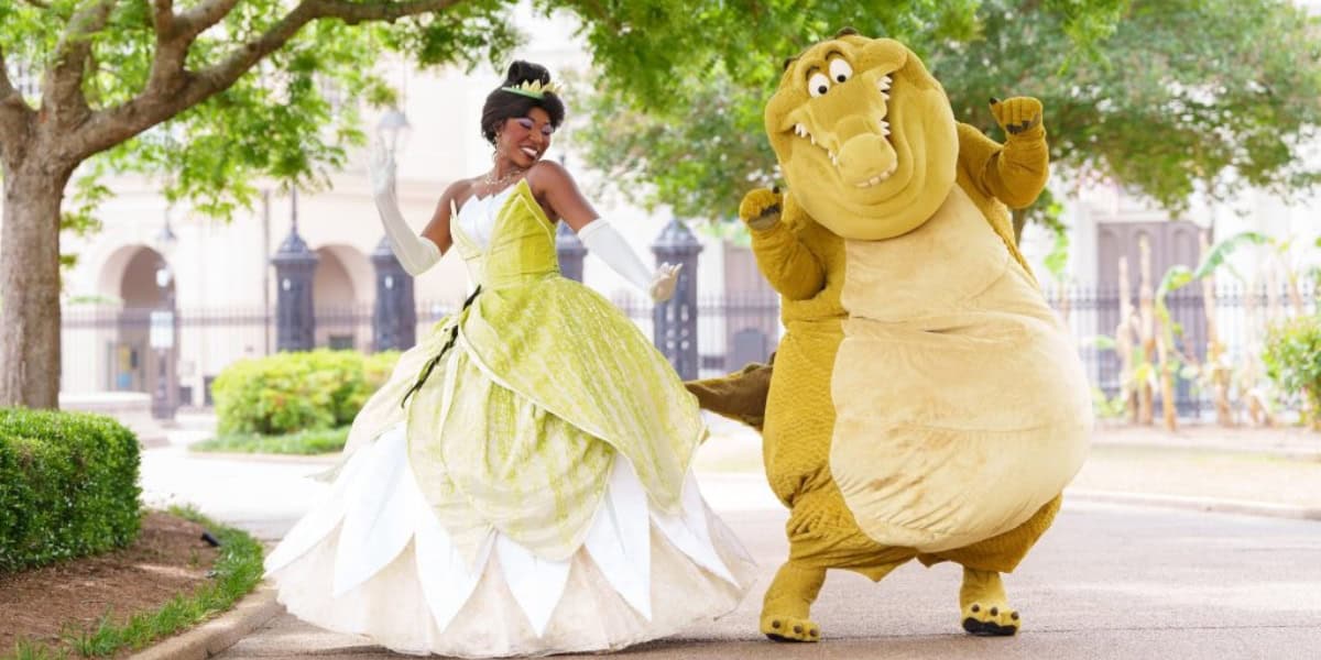 A person dressed as Princess Tiana in a green and white gown dances next to someone in a large, brown alligator costume. They are outdoors on a path with lush greenery and trees in the background, creating a joyful and playful scene.
