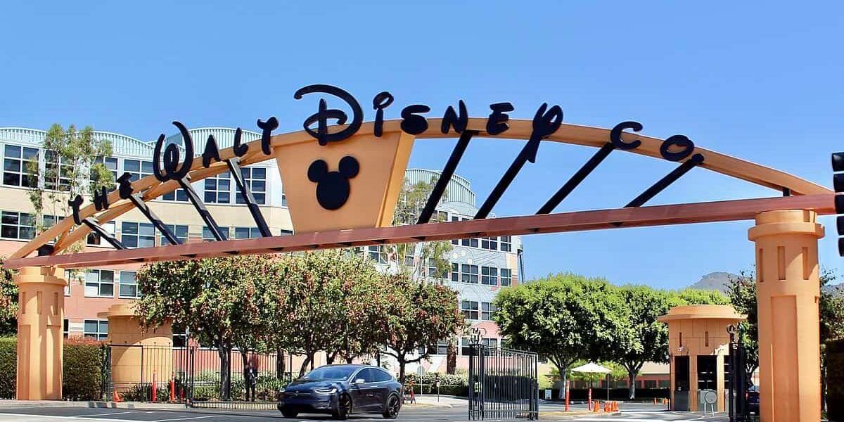 The iconic entrance gate, featuring the stylized logo of the Walt Disney Company with a Mickey Mouse silhouette, is set against a clear blue sky.