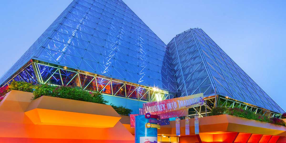 An image of two glass pyramidal buildings at sunset. The structures, part of EPCOT, are lit up with colorful lights. A banner reading "Journey Into Imagination" is displayed near the entrance of the building in the foreground. Trees and greenery surround the base of the pyramids, leading to a Disney Vacation Club Member Lounge.