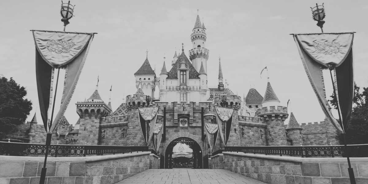 Black and white photo of a castle with tall spires, intricate stonework, and flags. An archway serves as the main entrance. Two banners bearing ornamental designs flank the pathway leading to the entrance. The sky above is cloudy and overcast, adding a touch of melancholy to what could be mistaken for a Disney Resort.