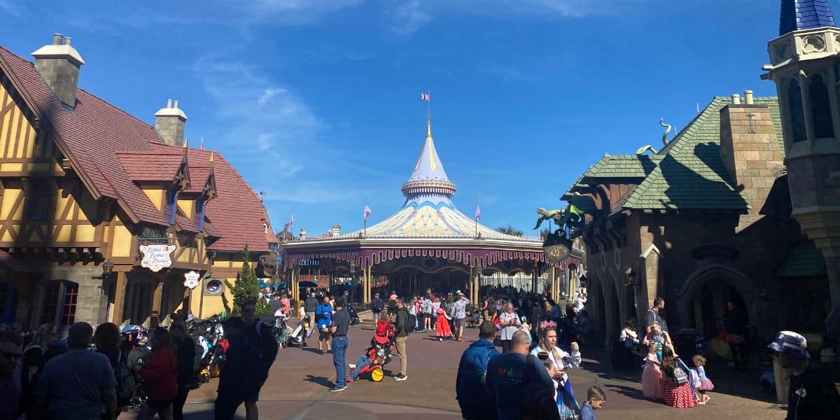A bustling theme park scene with people walking, pushing strollers, and taking photos. Colorful medieval-style buildings line the path, leading to a central carousel with a pointed roof. The sky is clear and blue, reminiscent of the magical charm found at Disney World.
