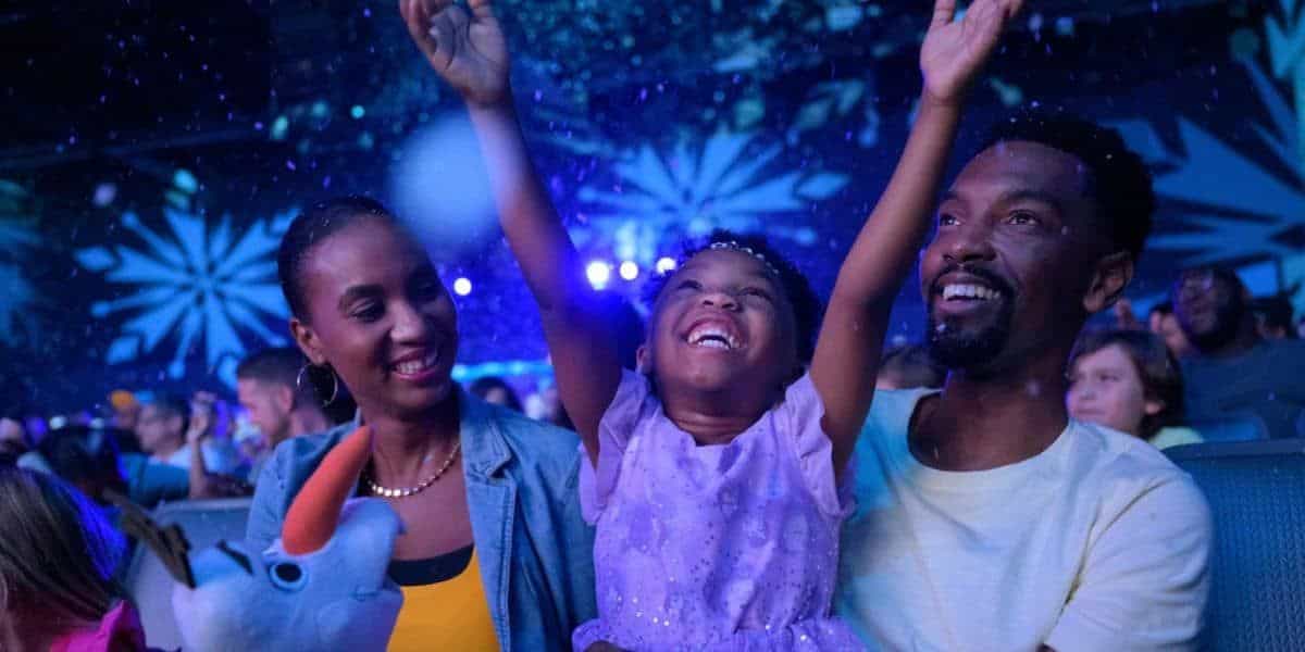 A family of three enjoys a night out at Disney Springs, having arrived via the Lynx Bus. The young girl in the middle, wearing a purple dress, joyfully raises her arms. Her parents sit on either side, smiling warmly. The dark background is illuminated by blue lights with an abstract pattern as the guests make their way to Epic Universe.