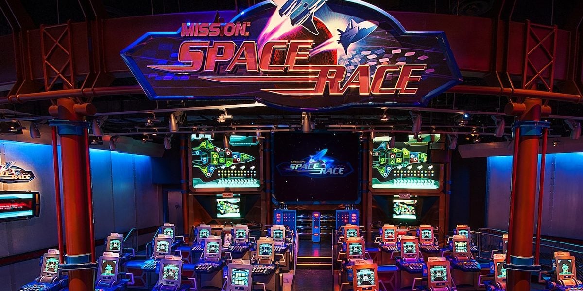 A futuristic space-themed arcade with rows of brightly lit gaming stations and a large "Mission: Space Race" sign above, featuring neon lights and a rocket graphic. Multiple screens display space visuals.