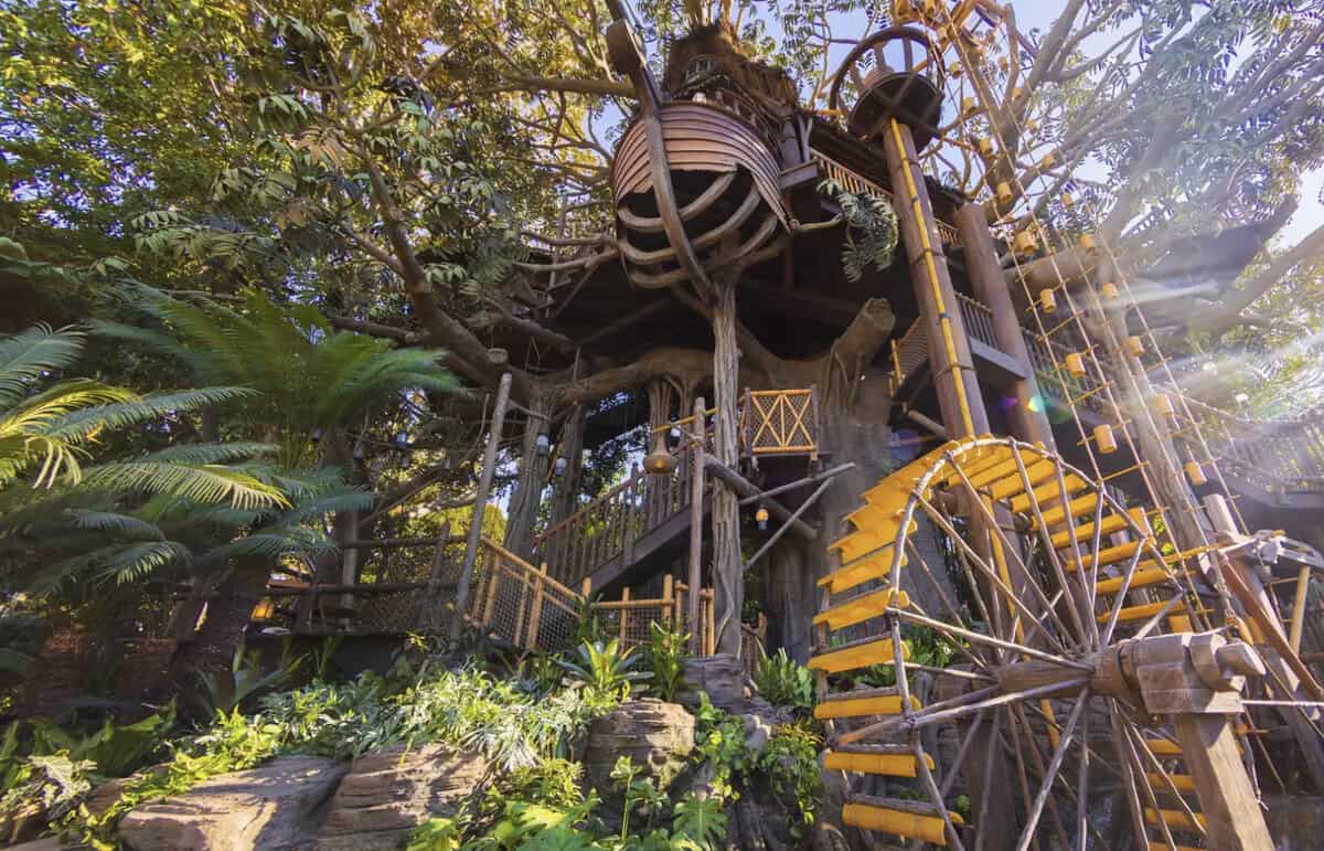 A whimsical treehouse with a water wheel beside it, surrounded by lush greenery and bathed in sunlight filtering through the trees.