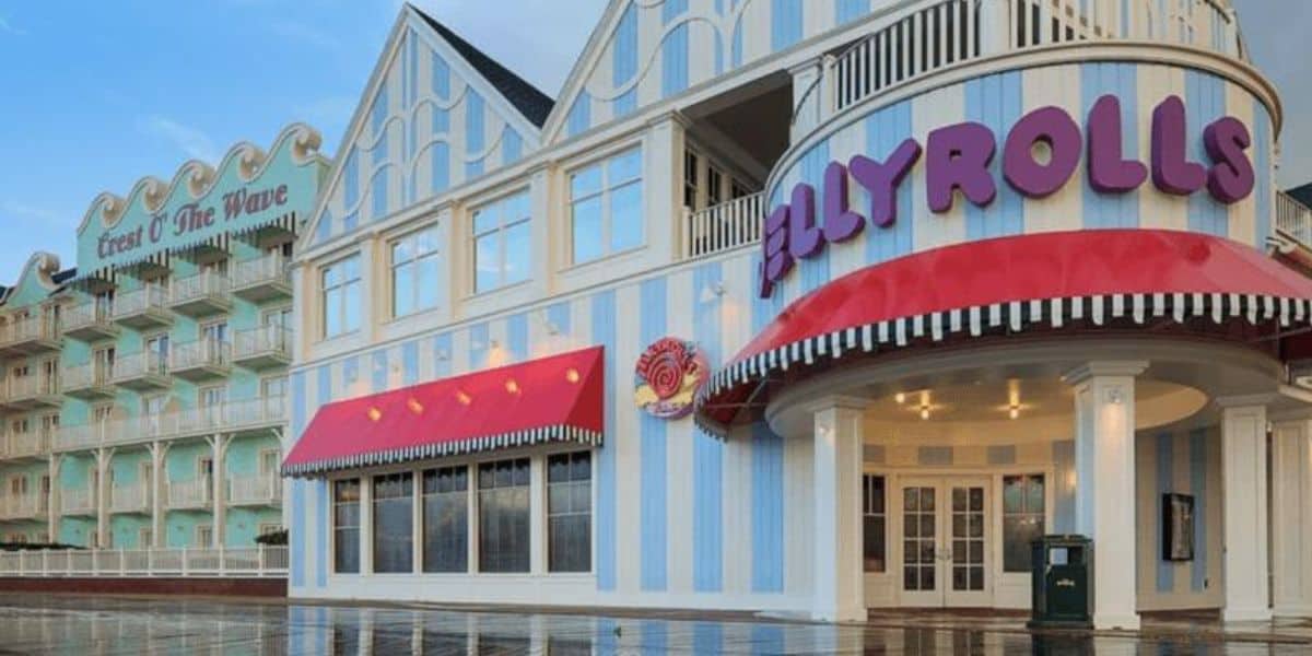 The exterior of Jellyrolls with pastel blue and white stripes catches the eye. A bright red canopy with the sign "Jellyrolls" in purple proudly marks its place at Walt Disney World. Nearby, another structure is labeled "Crest of the Wave," while the shiny ground glistens as if freshly kissed by rain.