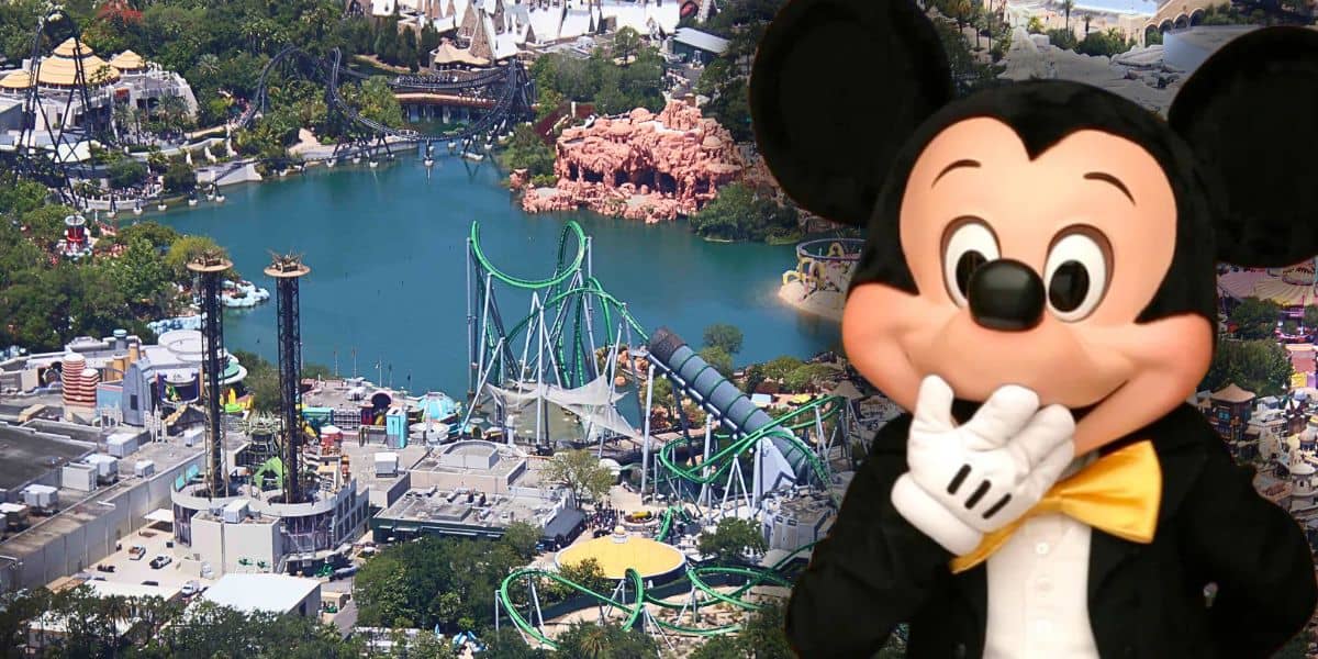 An aerial view of an amusement park featuring various rides and attractions around a lake, including roller coasters and themed buildings. In the foreground, a large costumed character resembling Universal's famous Despicable Me minion is playfully holding his gloved hand to his face.