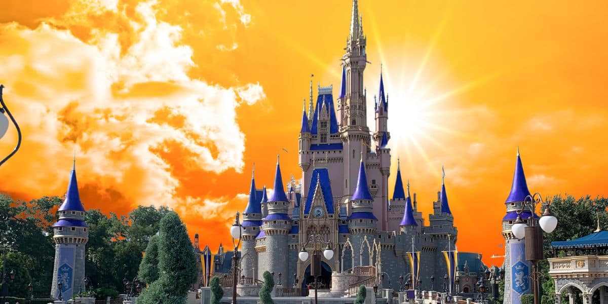 A stunning castle with blue and gold spires stands tall under a vibrant, orange sunset sky. The sun shines brightly, casting a warm glow over the scene. Trees and a neatly manicured garden frame the foreground, evoking a magical Disney-like charm.