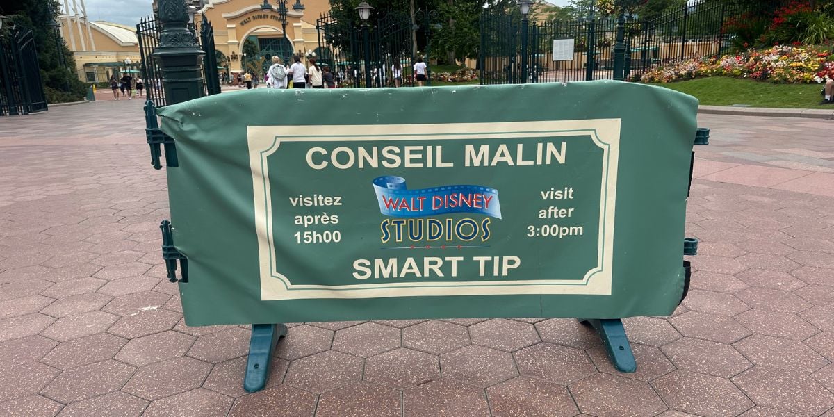 A green sign at Walt Disney Studios says "CONSEIL MALIN SMART TIP," advising visitors to visit after 3:00pm (15h00). The sign is in both French and English and is set on a paved walkway near the entrance.