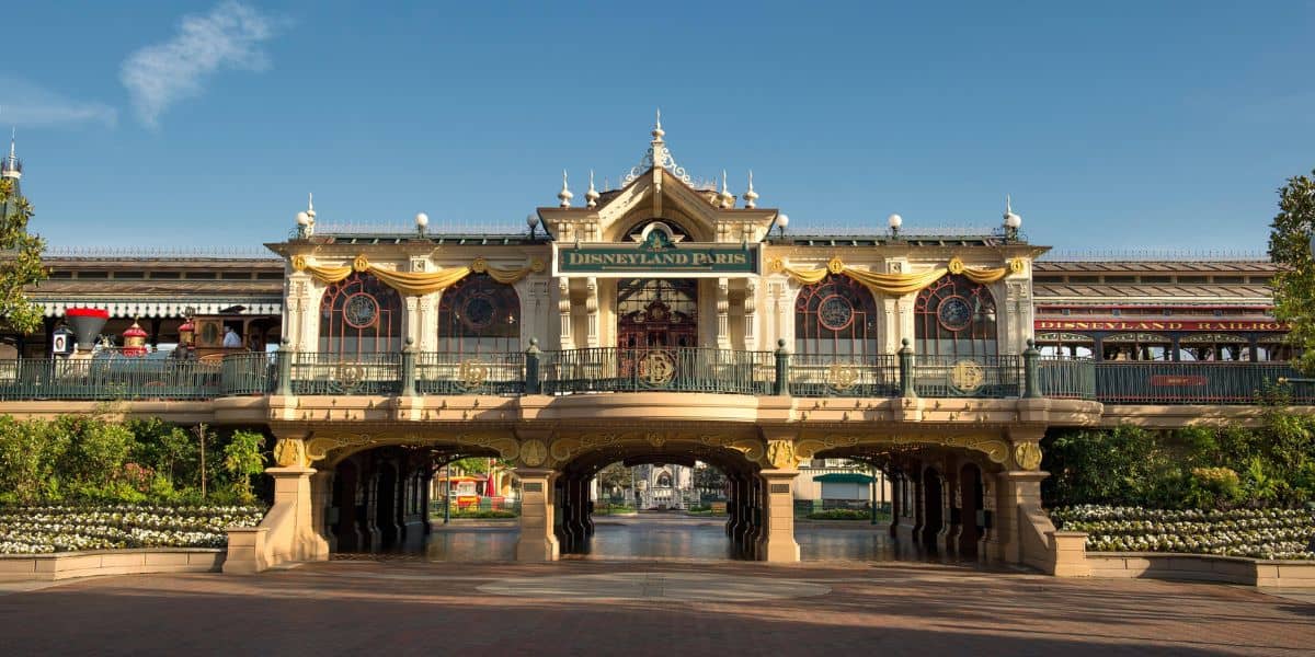 The image shows the main entrance of Disneyland Paris under a bright, clear sky. The building features ornate architecture with a sign reading "Disneyland Paris" and is decorated with gold-painted accents and floral arrangements. In the background is the Disneyland Railroad.