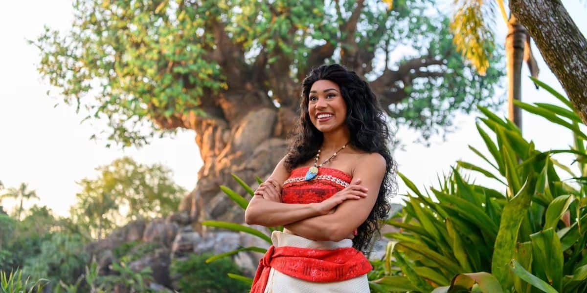 A woman dressed as moana stands with crossed arms, smiling, in front of the tree of life at a theme park, surrounded by lush greenery.