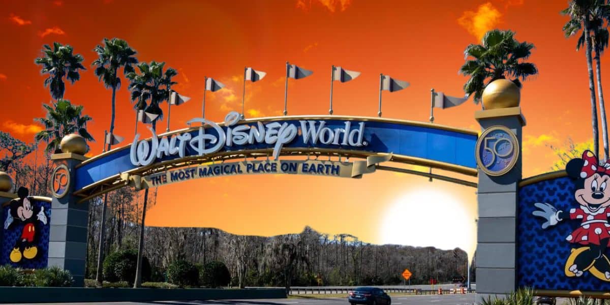 A grand entrance to Walt Disney World, featuring the iconic blue and gold archway with "The Most Magical Place On Earth" slogan. Mickey Mouse and Minnie Mouse flank each side. The sky glows a vibrant orange, suggesting either sunrise or sunset.