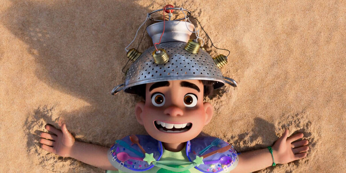 A smiling animated boy, Elio, lies on sandy ground with arms spread out. Wearing a colander hat with dangling light bulbs and a colorful, makeshift outfit, he looks cheerful and excited—ready for an Around the World Disney Parks adventure.
