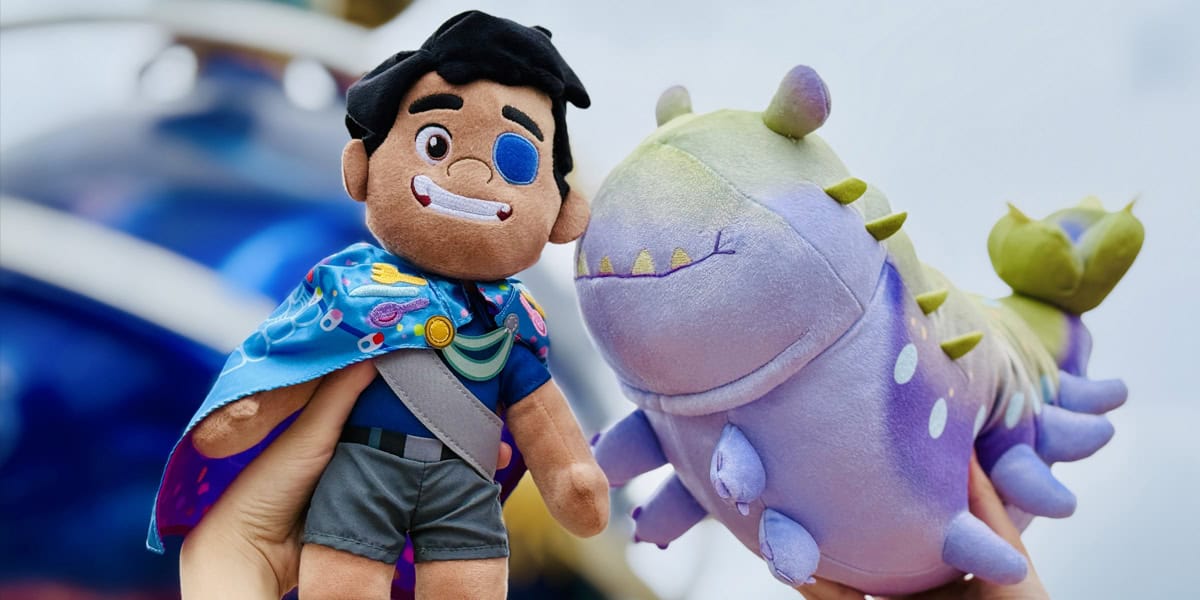 Two hands hold colorful plush toys: one is a smiling character with dark hair, an eyepatch, and a blue cape; the other is Elio, a pastel purple and green creature with small horns and a playful expression—perfect for Disney Parks fans around the world.