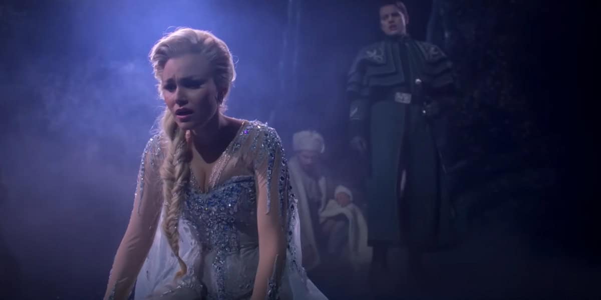 A woman in a sparkling light blue dress kneels on the ground, looking distressed—a scene reminiscent of Frozen The Broadway Musical. Behind her, a man in a dark coat stands as another figure sits in the misty, dimly lit background holding a baby.