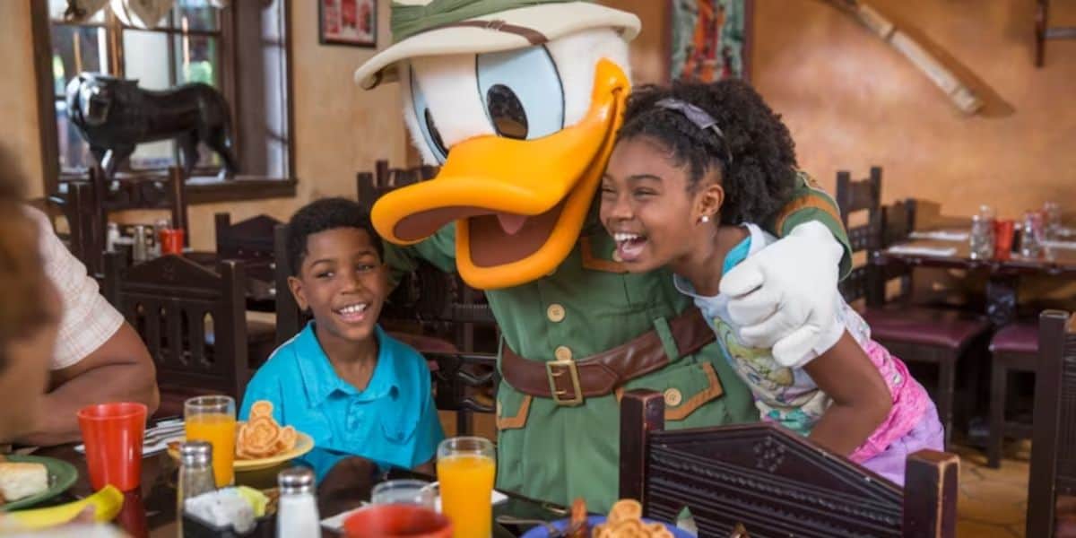 Two children are joyfully posing with a person dressed as Donald Duck in an explorer outfit at a restaurant. They are sitting at a table with waffles and drinks, enjoying the themed dining experience. The restaurant has a warm, rustic interior with various decorations.