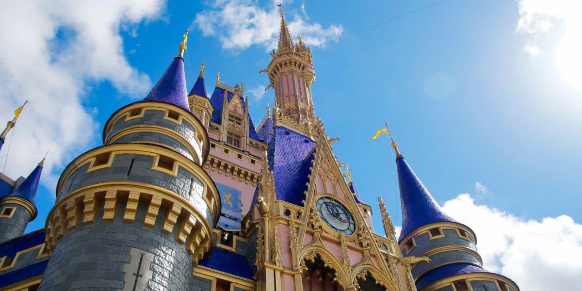 The Cinderella castle with blue spires and golden accents stands against a bright blue sky with scattered clouds at the heart of Walt Disney World Resort. The central spire is particularly tall, adorned with a flag at its peak, while a prominent clock greets visitors above the entrance inside Magic Kingdom expansion.