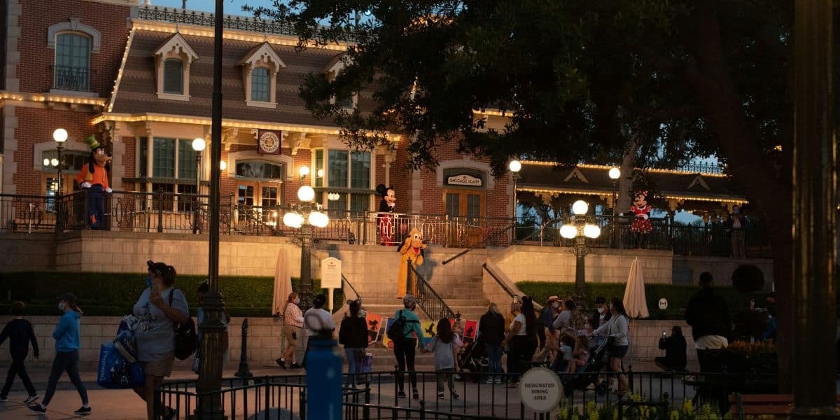 A lively scene unfolds in front of a well-lit, two-story brick building at dusk in a Disney theme park. The building features many windows and ornate detailing. People are walking and gathered around, with a character performer on the steps and several others interacting with guests.