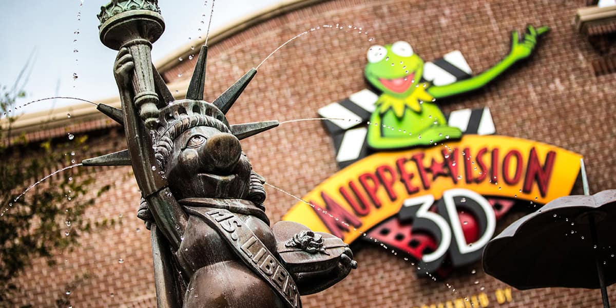 A whimsical fountain shaped like Miss Piggy as the Statue of Liberty sprays water, with the colorful Muppet*Vision 3D attraction sign and a large image of Kermit the Frog on a brick building at this playful Disney spot.