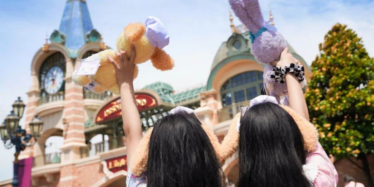 Two people with long dark hair stand with their backs to the camera, holding plush toys high in the air. The backdrop features a whimsical building with a clock tower, reminiscent of an amusement park entrance. The sky is clear and sunny.