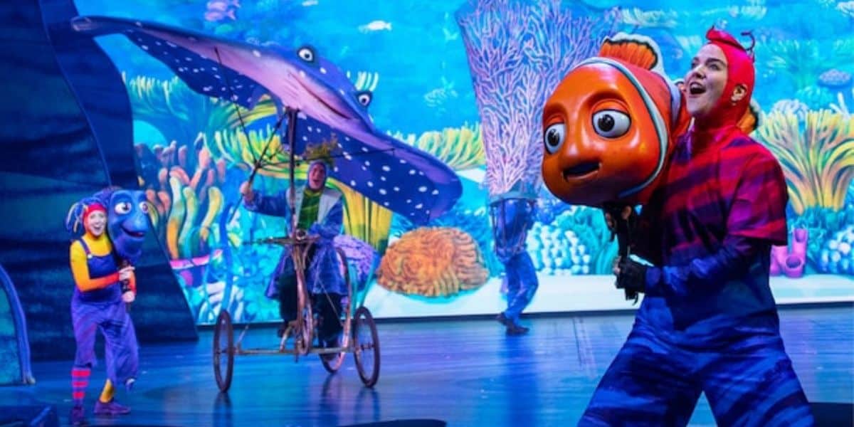 Performers in vibrant costumes on stage bring aquatic characters to life in a mesmerizing Finding Nemo Show. A person holds a large fish puppet resembling a clownfish, while another rides a bicycle with a stingray decoration. The colorful backdrop at Disney World depicts an enchanting underwater scene with coral.
