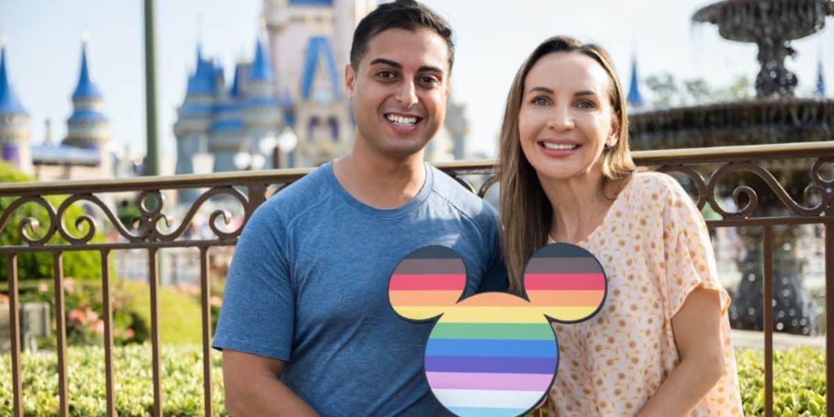 A smiling man and woman stand in front of a Disney World castle. The man holds a rainbow-colored Mickey Mouse head sign, celebrating a beloved gay icon. Both are casually dressed and appear happy in the bright, sunny setting. Metal railings and a fountain are also visible in the background.